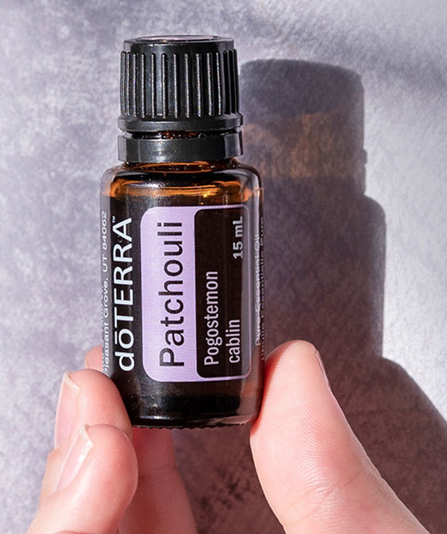 Рука держит флакон эфирного масла пачули doTERRA 15 мл на фоне светлой ткани с тенями, официальное фото doTERRA